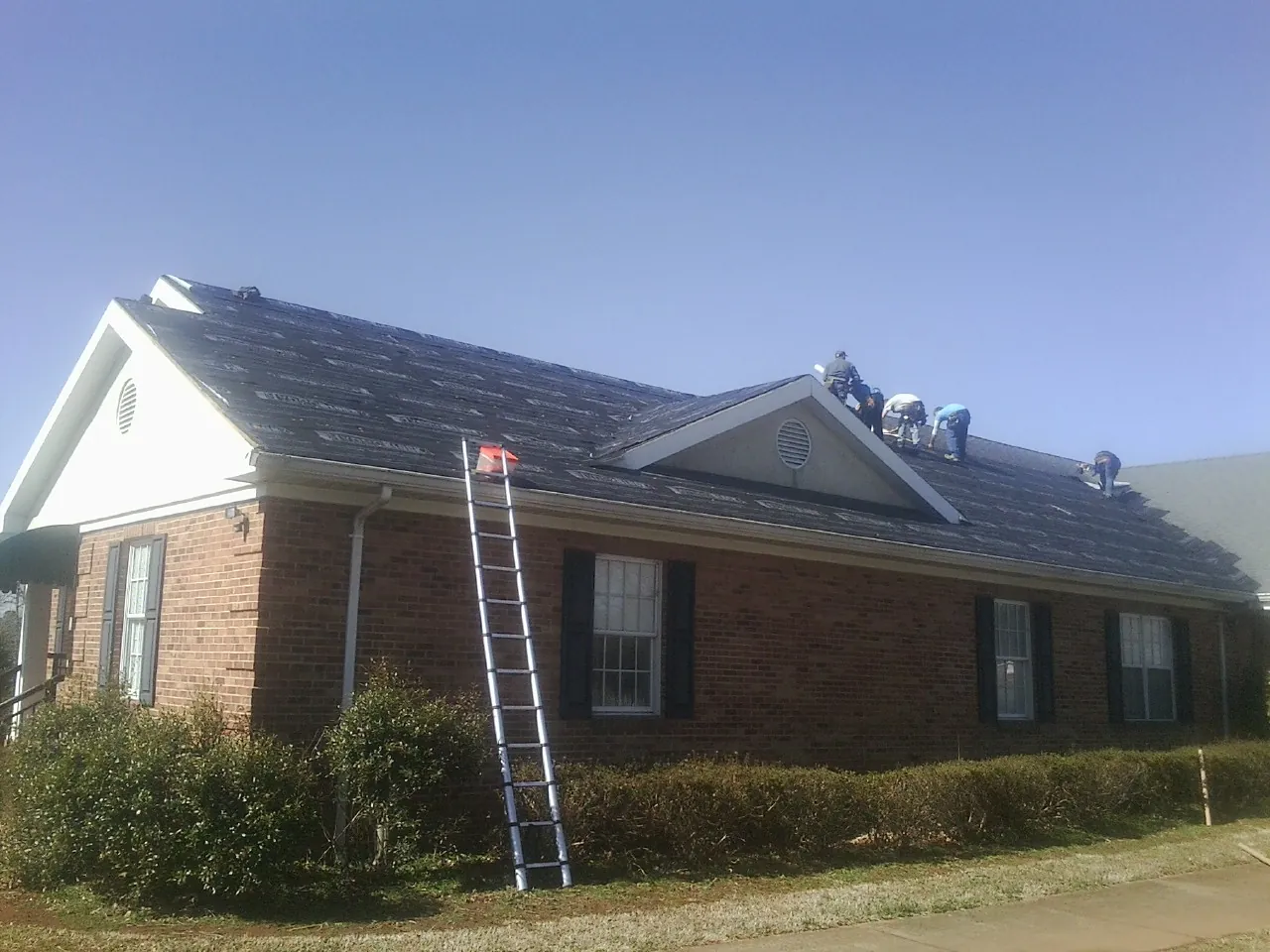 Crew tearing shingles off a brick ranch roof with a ladder set — Metal Roof Repair work by our crew in New Auburn