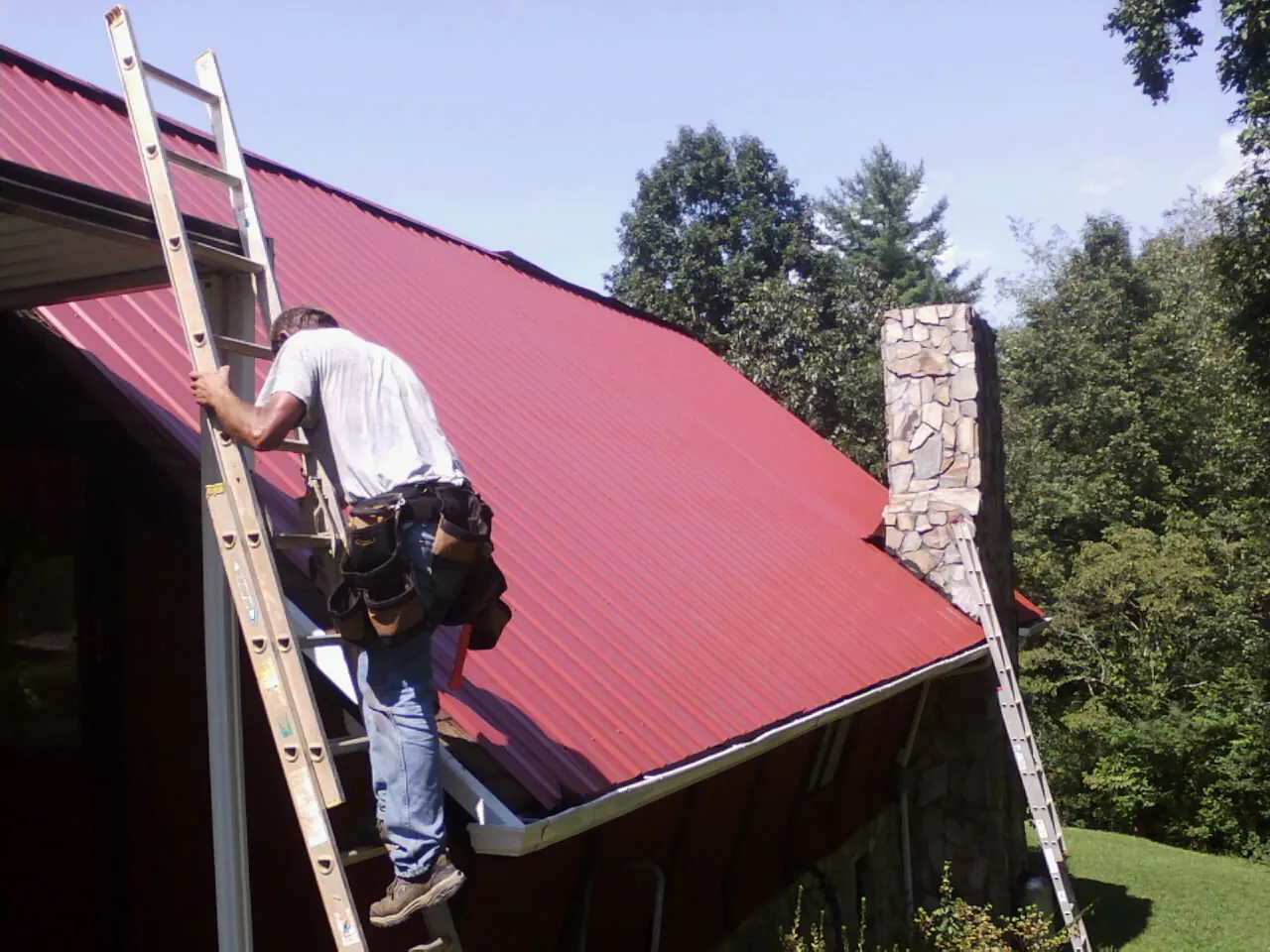Roofer on a ladder beside a red metal roof and stone chimney — Soffit & Fascia Repair by Roofing Brothers in New Auburn