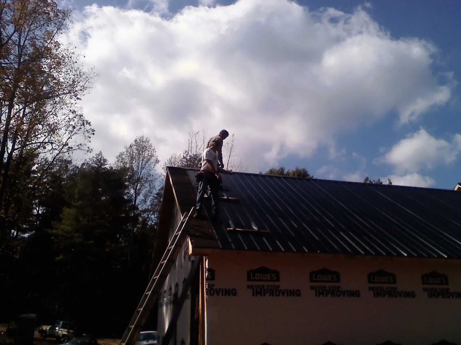 Roofer fastening dark metal panels high against a cloudy sky, from a Roof Inspection job in New Auburn